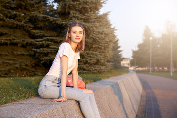 Photo of a beautiful blonde girl sitting on the railing in the Park at sunset. Woman resting on the street and waiting for friends after a work shift.