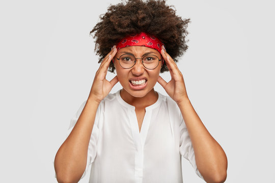 Stressful black girl has terrible headache, touches head with hands, clenches teeth, has Afro hairstyle, cant concentrate on something, poses against white background. Negative feeling concept