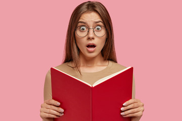 Stunned surprised teacher with book in hands, looks at pupil who made mess in classroom, wears transparent glasses, keeps jaw dropped, poses against pink background. Clever astonished student