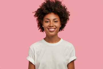 Indoor shot of attractive dark skinned girl with toothy smile, shows white perfect teeth, being in high spirit, has date with boyfriend, looks directly at camera, isolated on pink background.