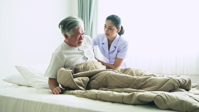 Senior man in bed trying to get up and nurse helping him. Old asian man and beautiful asian nurse woman in bedroom and open curtain. Senior home service concept. Side shot.