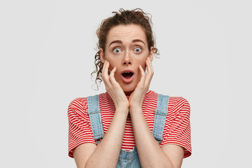 Bugged eyed terrifc girlfriend with curly hair, freckled skin, opens mouth widely, dressed in casual t shirt, denim overalls, stands against white background, being shocked with latest news.