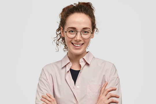 Attractive European girl with charming smile, keeps arms folded, wears round spectacles, being in good mood, expresses happiness, poses against white background. Freckled smiling teenager indoor