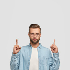 Photo of unshaven hipster dressed in casual shirt, points with both index fingers upwards. shows...