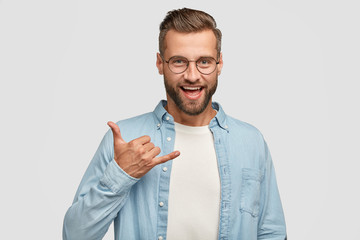Friendly looking positive hipster with dark stubble, gestures indoor, shows shaka sign, being in...