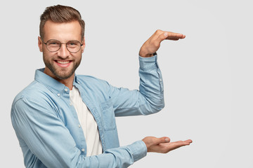 Cheerful unshaven guy stands sideways, holds background, gestures with both hands as if carrying something, shows height of thing, dressed in fashionable clothes, isolated over white background