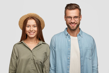 Isolated shot of happy friends enjoy pastime together, have toothy smiles, rejoice positive moments, stand closely against white studio wall. Glad woman in straw hat and her boyfriend have date