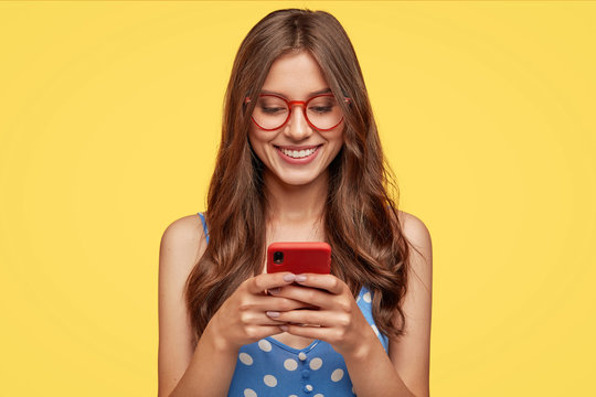 Content teenager with long hair, holds modern cell phone, scrolls through social networks, has cheerful expression, wears spectacles and casual dress, isolated over yellow background, recieves message