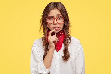 Embarrassed hipster or female teenager has displeased facial expression, keeps index finger near lips, looks with frustration, sees no way out dressed in fashionable clothes stands against yellow wall