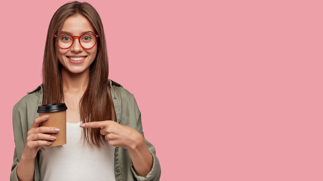 Drink this! Cheerful beautiful European woman in spectacles, holds disposable cup of coffee, indicates at beverage, poses against pink background with free space aside for your promotional content