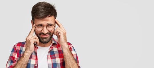 Frustrated man has dark bristle, keeps index fingers on temples, focuses on something important, looks with displeased expression aside, isolated over white background with blank copy space.