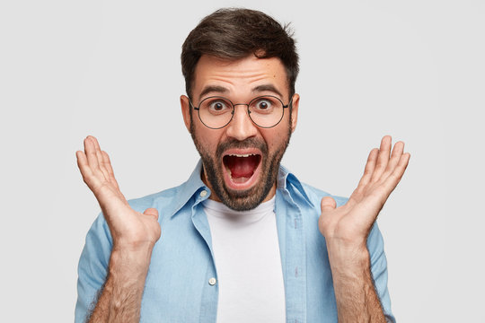 Surprised young amazed man with stubble opens mouth widely, clasps hands near face, wears glasses for good vision, hears unexpected positive news, isolated over white background. Emotions concept