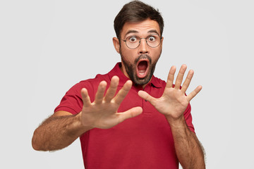 Body language concept. Frightened man with fearful expression, gestures with palms, makes defense sign, exlaims with astonished expression, dressed in red casual t shirt isolated over white background