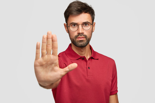 Concerned serious bearded man in round eyewear pulls palm towards camera, stops or warns you from making bad choice, shows enough or rejection gesture, isolated on white wall. Slow down, please!