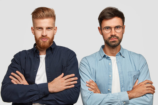 Horizontal shot of serious self assured guys listen something attentively, stand closely, keep hands crossed over chest, dressed in fashionable shirt, stand together against white background