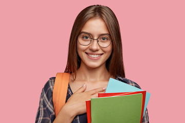 Thankful brunette woman with gentle smile, keeps hand on chest, expresses good feeling to friends and relatives, pleased to hear positive results of entrance exam carries bag and scientific literature