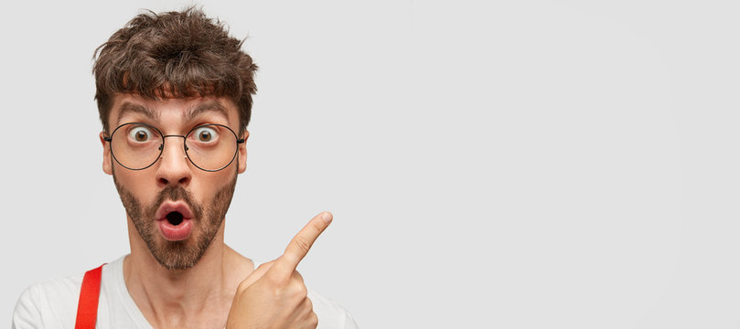 Emotive hipster with surprised expression, gasps from wonder, indicates at copy space with index finger, shows free space for your promotion, wears round transparent glasses. Advertisement concept