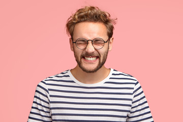 Obraz premium Photo of attractive cheerful man laughs positively at camera, keeps eyes closed, dressed in casual striped t shirt, round spectacles, models in studio against pink background. Happy hipster indoor