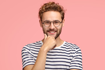 Horizontal shot of satisfied self assured guy with pleasant smile, keeps hand on chin, wears casual clothes, rejoices good resuls or achievement, stands against pink background. Happiness concept