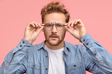 Horizontal shot of pleasant looking guy looks scrupulously through glasses, tries to notice...