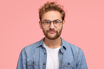 Obraz premium Funny man makes grimace, pouts lips, has comic facial expression, curly hair, bristle, listens some information, dressed in fashionable outfit, isolated over pink background. People, lifestyle concept