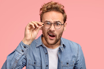 Fototapeta na wymiar Indignant young Caucasian guy looks desperately at camera, wears spectacles, opens mouth, has surprised gaze, expresses frustration, wears round spectacles, stands against pink studio background