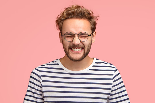 Photo of attractive cheerful man laughs positively at camera, keeps eyes closed, dressed in casual striped t shirt, round spectacles, models in studio against pink background. Happy hipster indoor