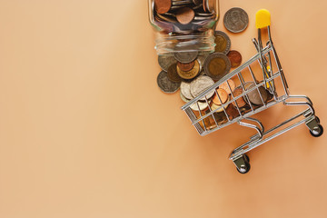 Fototapeta na wymiar Money and coins form glass jar to mini shopping cart or trolley on beige color background for spending plan, investment, business and finance concept
