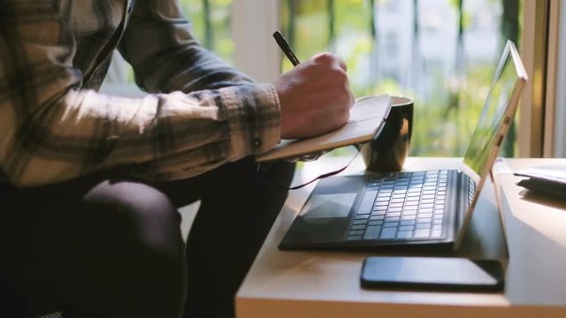 Medium shot of man working on laptop at home office, home scenery of employee sitting on couch and using notepad calendar, window in background, shot in 4K UHD