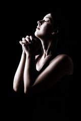Faithful woman praying, hands folded in worship to god with head up and closed eyes in religious fervor, on a black background. Concept for religion, faith, prayer and spirituality. - obrazy, fototapety, plakaty