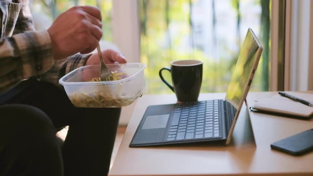 Man workaholic eating pasta from a plastic lunch box while he is working on laptop at home office, no break, static shot of employee sitting on couch, 4K UKD