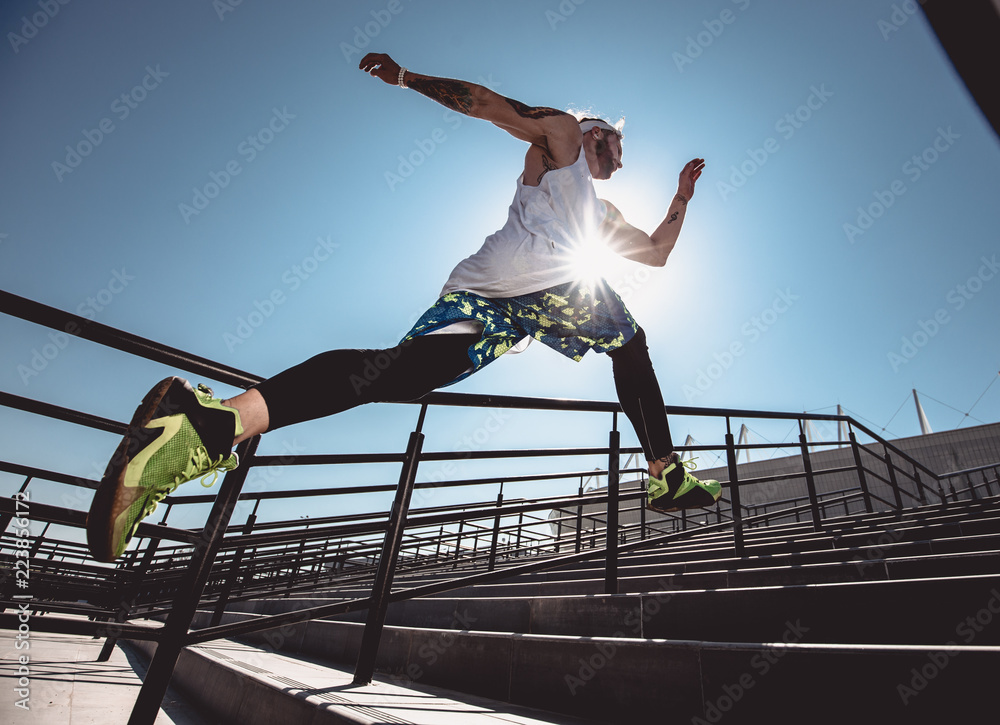 Wall mural Handsome young muscular man in modern sport clothing run up the stairs outdoor at bright sunny day. Wide angle photo of a jogging man. Sport lifestyle