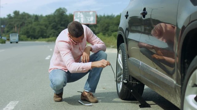 Handsome millennial male driver lifting his car on the jack to change flat tire on the road after tyre puncture during travel. Man with jack under his car lifting vehicle to replace tires on roadside.
