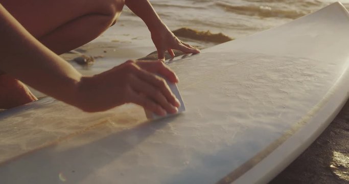 Close up shot of surfer girl waxing surfboard on beach at sunset 