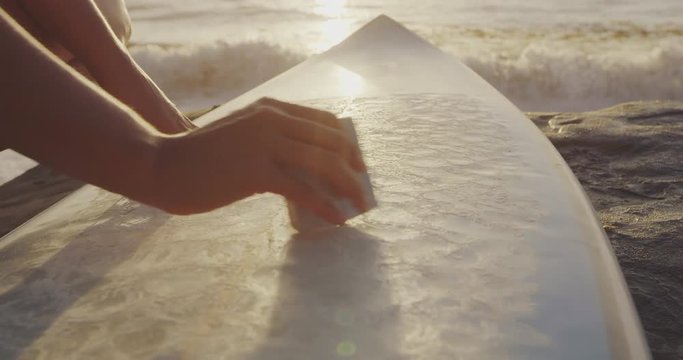 Close up shot of surfer girl waxing surfboard on beach at sunset 