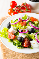 Fresh Greek salad made of cherry tomato, ruccola, arugula, feta, olives, cucumbers, onion and spices. Caesar salad in a white bowl on wooden background. Healthy organic diet food concept.