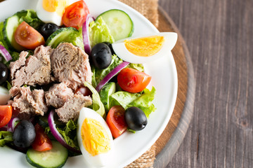 Fresh fish tuna salad made of tomato, ruccola, tuna, eggs, arugula, crackers and spices. Caesar salad in a white bowl on wooden background