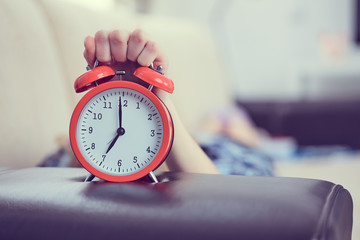 Young girl lies on the couch and stretches her hand to the red alarm clock to turn it off. Late wake up.