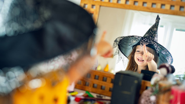 Adorable young girl wearing witch hat looking at herself in the mirror, pointing with finger and laughing. Happy child in halloween costume looking in the mirror.