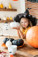 Obraz premium Cute little girl in witch costume sitting behind a table in Halloween theme decorated room, holding hand painted pumpkin and smiling. Halloween holiday concept.