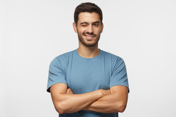 Handsome young man in blue t-shirt, with crossed arms smiling and winking, looking at camera isolated on gray background - obrazy, fototapety, plakaty