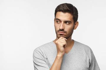 Close up portrait of young man looking left, holding chin as if thinking of something, trying to...