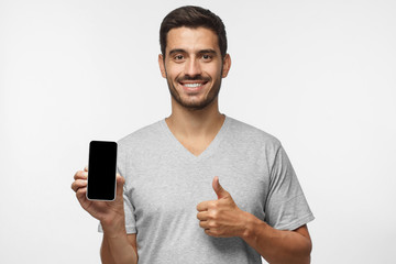 Attractive happy young man holding blank smartphone, smiling at camera, showing thubms up gesture,  isolated on gray background