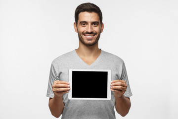 Young man isolated on gray background holding tablet and showing blank screen with happy smile as...