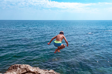 A boy is jumping from the cliff into the sea on a hot summer day. Holidays on the beach. The concept of active tourism and recreation