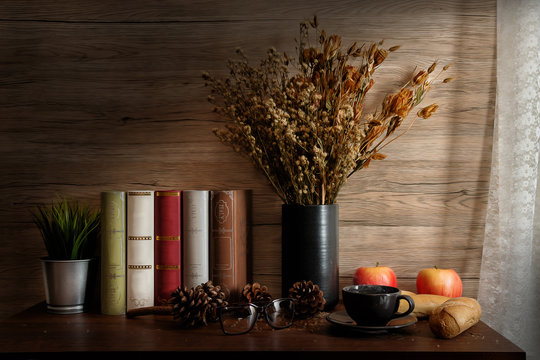 Still life of breakfast set of tea and bread and apple on wooden table with old stlye eye glasses and book and a vase of dry flowers
