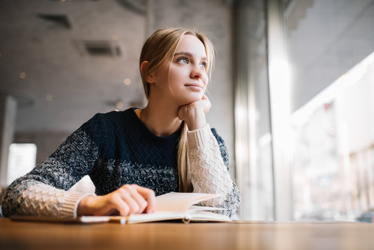 Young attractive university student studying at modern library. Cute caucasian woman with beautiful blue eyes sitting in loft cafe, planning working process, thinking about project. Exam preparation