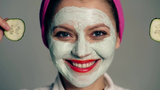 Close up of a girl with a face mask and a towel on her head that closes her eyes with cucumbers on a gray background. Face care concept. Beautiful girl with cosmetic mask on her face.