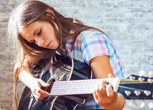 portrait of young teenager brunette girl with long hair playing an black acoustic guitar on gray wall background