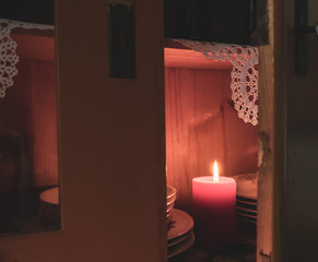 View into an open doors of a vintage cupboard illuminated with a lit red candle in a dark room, with old dishes, apples and crocheted doily. Autumn composition or happy holiday season background.
